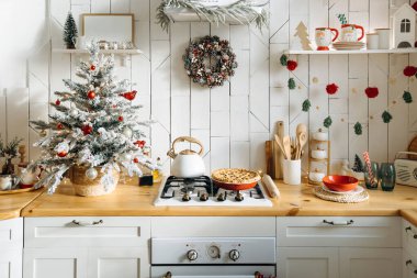 Küçük buzlu bir Noel ağacı, bir kozalak çelengi, kırmızı ve beyaz süsler ve tezgahın üzerinde taze pişmiş bir turta bulunduran şenlikli mutfak, sıcak bir tatil ortamı yaratıyor..
