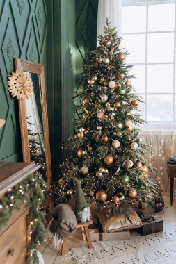Güzel dekore edilmiş, yeşil, altın, bakır ve gümüş takılarla süslenmiş Noel ağacı bir pencerenin yanında duruyor. Yanındaki ahşap çerçeveli ayna ağacı yansıtıyor, hediyeler ve yerde bir tatil cücesi var..