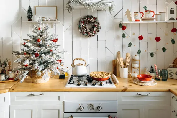 Küçük buzlu bir Noel ağacı, bir kozalak çelengi, kırmızı ve beyaz süsler ve tezgahın üzerinde taze pişmiş bir turta bulunduran şenlikli mutfak, sıcak bir tatil ortamı yaratıyor..