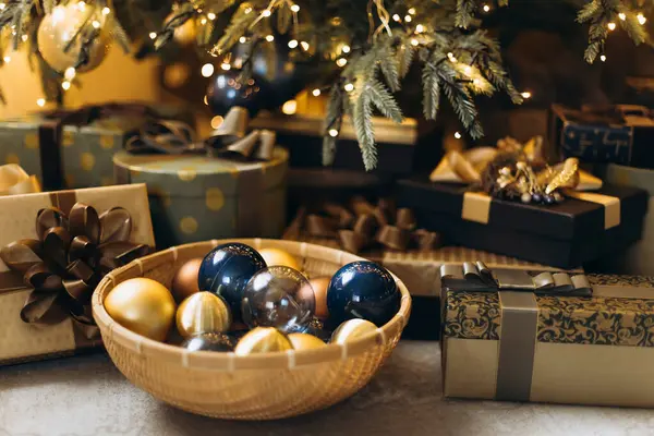 Altın ve lacivert Noel süsleriyle dolu kase yakın plan görüntüsü, kurdeleli ve fiyonklu hediyeler, arka planda ışıklandırmalı Noel ağacı, şenlikli atmosfer, sığ DOF.