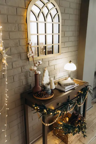 Dekoratif aynalı bir sahne, aydınlatılmış masa lambası, kitaplar, vazodaki pamuk dalı, ve sicim ışıkları ve çam ağacından çelenklerle süslenmiş bir masanın üzerindeki Noel süsleri..