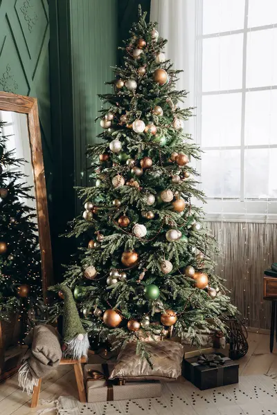 Altın, gümüş ve yeşil süslerle süslenmiş uzun yeşil Noel ağacı büyük pencere ve aynanın yanında duruyor. Pofuduk cüce, battaniye ve hediyeler yerde oturur ve neşeli bir ortam yaratır..