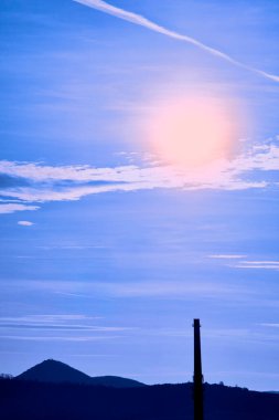 Mountains or hills, factory chimney on sunny background, blue sky with white clouds, contrail, and yellow shining sun