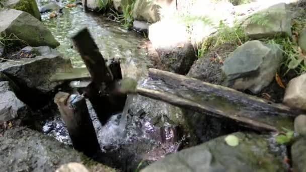 在一条天然的小溪流中旋转着的水轮 在石头之间流过的河流上 一个木制渠道的水流驱动的小型水磨机在工作 向儿童展示绿色技术 — 图库视频影像