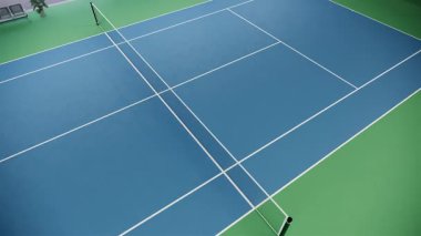 Mavi ve yeşil yüzeyi ve kıvrımlı çatı altında beyaz çizgileri olan boş bir tenis kortu