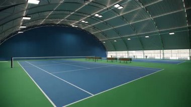 Mavi ve yeşil yüzeyi ve kıvrımlı çatı altında beyaz çizgileri olan boş bir tenis kortu