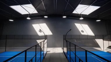 Hava aracı görüntüsü, spor tesisi bölgesinde bulunan bir depo binasının içindeki kullanılmayan yeşil ve mavi padel tenis kortlarını yakınlaştırıyor. Gündüz olmasına rağmen ışıklar açık.