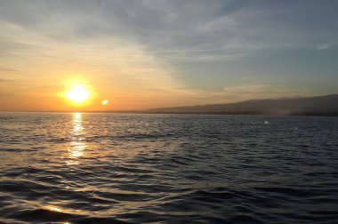 Endonezya, Bali 'deki sakin okyanusun üzerinde güneşin nefes kesici manzarası, dağ siluetleri ve altın ışık yansımaları.