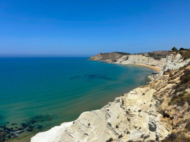 Turkuaz deniz, altın kumlu plaj ve dramatik beyaz uçurumlarla çarpıcı bir sahil manzarası. Gökyüzü açık ve mavi, ve kıyı şeridi zarifçe kıvrılıyor, huzurlu, resimsel bir manzara sunuyor..