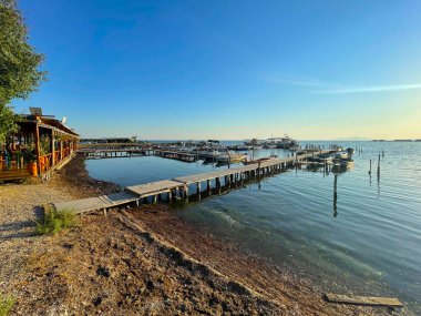 GREECE.05.07.2025 balıkçı tekneli küçük bir marinaya çıkan ahşap bir rıhtım. Sakin, berrak su mavi gökyüzünü yansıtır. Huzurlu kıyı sahili uzak adalar ve sakin bir limanı içeriyor.
