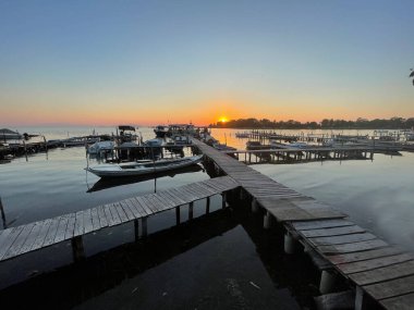 Limanda sakin sularda yüzen teknelerle huzurlu bir günbatımı. Ahşap iskele, gözü güneşin battığı ufka götürüyor, sıcak turuncu ve mor renklerde gökyüzünü boyuyor..