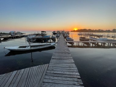 Limanda sakin sularda yüzen teknelerle huzurlu bir günbatımı. Ahşap iskele, gözü güneşin battığı ufka götürüyor, sıcak turuncu ve mor renklerde gökyüzünü boyuyor..