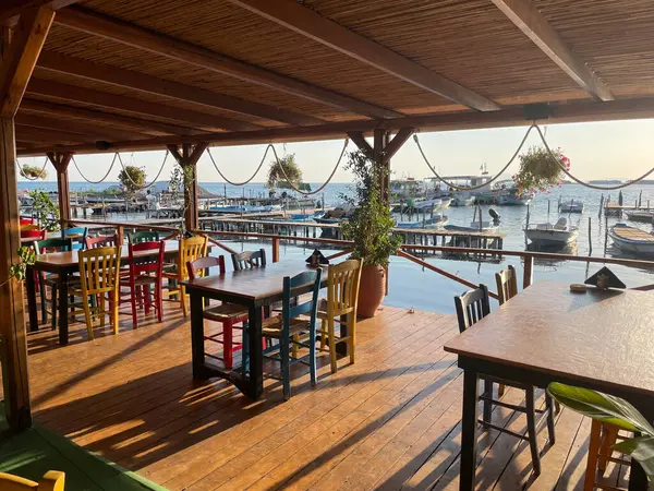 Renkli sandalyeleri ve ahşap masaları olan, gün batımında tekneleri olan huzurlu bir marinaya bakan samimi bir sahil restoranı terası. Mükemmel bir tatil ve seyahat sahnesi, sıcaklık ve cazibeyle dolu..
