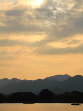 Gün batımında, dağların ve ayın arka planında sakin denizde yüzen bir siluet tekne. Altın ve sarı renk. Seyahat, geçmiş ve blog gibi şeyler için. Kavramları. Batı Gölü. Hangzhou şehrinin popüler parkı.