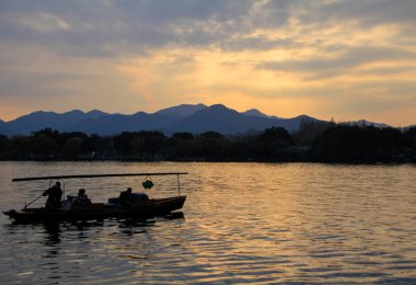 Bir siluet tekne ve bir adam gün batımında sakin denizde yelken açıyorlar. Dağların ve güneş ışınlarının arka planında. Dramatik gökyüzü ve bulut manzarası. Seyahat, geçmiş ve blog gibi şeyler için. Kavramları. Batı Gölü. Hangzhou şehrinin popüler parkı.