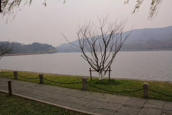 The path along the lake with bare trees. Foggy and calm seascape with mountains in the background. West Lake. Popular park of Hangzhou city China
