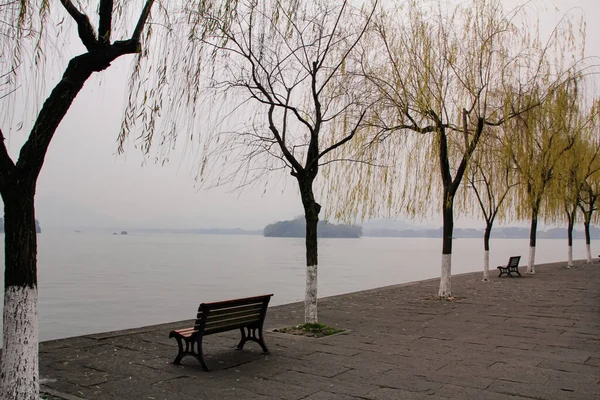 The path along the lake with bare trees. Foggy and calm seascape with mountains in the background. West Lake. Popular park of Hangzhou city China