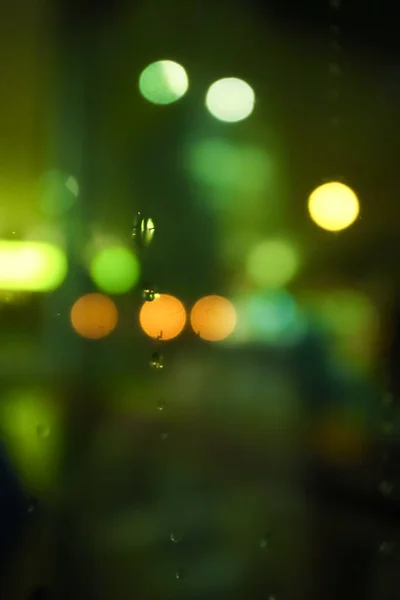 Bokeh effect of street lighting at night in green and yellow color with the focus of some raindrops as foreground. Looking through from car window.