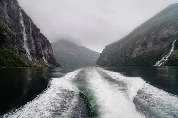 Norveç 'te bir fiyortta yelken açan bir teknenin dalgaları ve yamaçlarında yüksek kayalıklar.