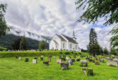 Norveç 'te ahşap bir kilise, yanında mezarlık ve bulutlu yeşil çimenler.