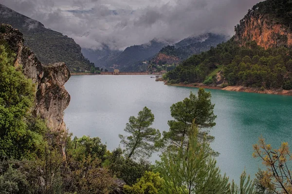 Dağlarla çevrili su deposu ve İspanya 'da bulutlu gökyüzü ve resimde çam ağaçları var.