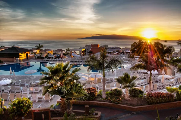 İspanya 'nın Tenerife Adası' nda gün batımında palmiye ağaçları ve yüzme havuzunun olduğu bir otel manzarası ve arka planda La Gomera volkanik adası.