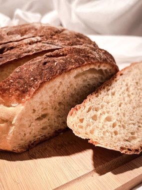 Taze pişmiş yuvarlak ekmek beyaz bir kumaş zemin üzerinde tahta bir tahta üzerinde dilimler halinde kesilmiş. Kapat..