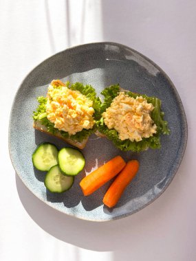 Beyaz arka planda gölgeli bir tabakta salatalıklı ve havuçlu yumurta salatalı sandviç. Sağlıklı kahvaltı.