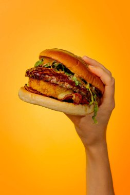 Turuncu bir arka planda parlak sulu bir burger tutuyor.
