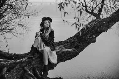 Young pensive woman in fedora hat sitting alone on fallen tree trunk by river and looking away. Black and white mindful female lost in thoughts. Monochrome art photo with fall melancholy mood concept.