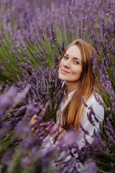 Woman sitting among the rows of purple lavender in blooming field. Portrait of female holding bouquet in full bloom purple lavender field. Girl with cheek dimples enjoying summer flower field.