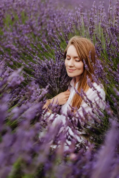 Young woman sitting among the rows of purple lavender in blooming field. Portrait of female holding bouquet in full bloom purple lavender field. Girl with cheek dimples enjoying summer flower field.
