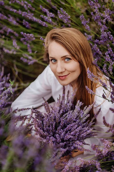 Woman sitting among the rows of purple lavender in blooming field. Portrait of female holding bouquet in full bloom purple lavender field. Girl with cheek dimples enjoying summer flower field.