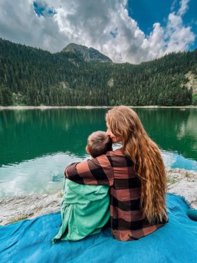 Durmitor Ulusal Parkı 'nda Kara Göl' ün kenarında oturan genç bir anne oğluna sarılıp öpüyor. Anne ve çocuk su kenarında piknik battaniyesinde dinleniyorlar. Mutlu aile doğada birlikte vakit geçiriyor..