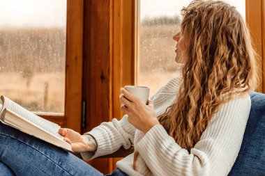 Kitap okuyan ve terasta kahve veya kakao içen genç bir kadın. Güzel bir kadın, bir fincan çayla koltukta oturup pencereden yağmuru izlerken heyecan verici romanlar okumaya tutkulu..