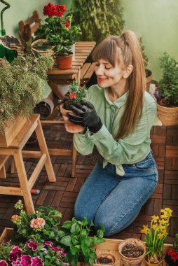 Mutlu kadın bahçıvan teras bahçesinde bitki dikiyor. Bahçıvan eldivenli bir kadın tahta kutunun önünde diz çöküyor ve elinde kil kapta karanfil çiçeği tutuyor. Balkonda bahçıvanlık..