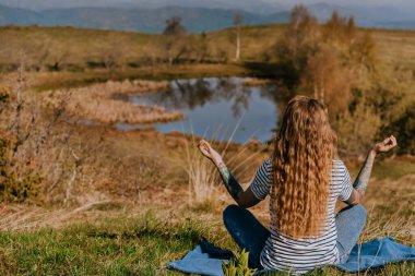 Young woman meditating sitting alone on hill near lake Nebeska Suza or Okruglica on Golija mountain, Serbia. Female solo traveler doing morning Yoga practice, enjoying silence and alone time outdoors. clipart