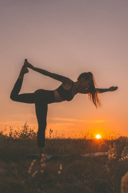 Gün batımında yoga yapan yogi kadının silueti. Yogayı seven bir kadın, Asya 'da tek ayak üstünde egzersiz minderinin üzerinde duruyor ve dengede duruyor. Rahatlama, vücut sağlığı ve açık hava fitness konsepti.