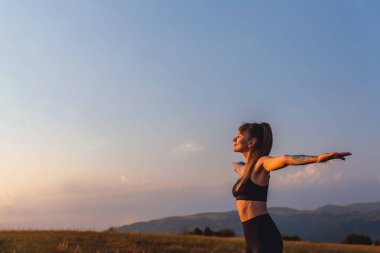 Yogi kadın yoga yapıyor gözleri kapalı duruyor ve gün batımında dağlarda kollarını açıyor. Mutlu ve özgür kadın nefes alma egzersizleri yapıyor gün batımının tadını çıkarıyor açık havada spor faaliyetlerinin ardından..