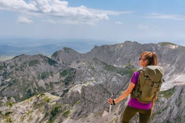 Female solo traveler with hiking backpack standing on rocky mountain peak and looking away. Woman on a summit of Bobotov kuk after successful climbing. Travel lifestyle and hiking adventure concept. clipart