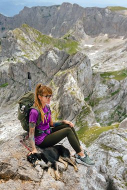 Female hiker sits with dog at Bobotov kuk mountain top. Woman hiker sitting on rock with relaxing mood and watching beautiful view. Travel with pet, outdoor activities and adventurous travel concept. clipart