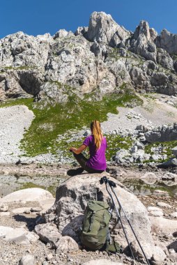 Back view of woman hiker sitting by mountain lake and resting, her trekking gear leaning against rock. Female traveling alone in mountains, relaxing during break on hiking trail in Durmitor park. clipart