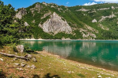 Piva Gölü 'nün büyük manzarasında küçük bir adam. Yaz günü Piva nehri kanyonunda mayo giymiş bir kadın turkuaz gölün kenarında duruyor. Balkanlar 'daki Durmitor dağlarının eteklerindeki Piva Doğa Parkı.