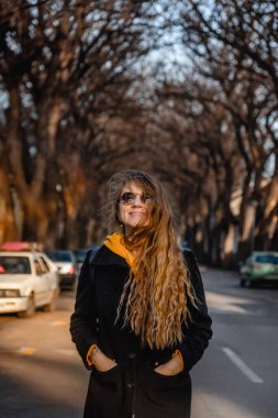 Happy woman walking in autumn city street in Kikinda. Female with long curly hair and dimples on cheeks wearing casual clothes enjoying sunny fall day in one of the most beautiful streets in Europe. clipart