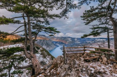 Banjska stena viewpoint with panoramic view of Drina river Canyon and lake Perucac on winter morning. Popular touristic observation deck in Serbia with wooden bench and a safety fence in wintertime. clipart