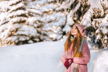 Gülümseyen kadın kar ormanlarında yürüyor, temiz hava soluyor, dondurucu güneşli günün tadını çıkarıyor. Kadın, karlı ormanda doğaya ve tatil haftasonuna hayran. Açık hava kış aktivitesi, doğa konseptinde yürüyüş..