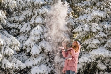 Kar ormanlarında eğlenen genç kadınlar. Sıcak atkı ve pembe ceketli bir kadın kar yağdırıyor, karlı köknar ağaçlarının arasında ilk karın tadını çıkarıyor. Mutlu kış ve Noel tatilleri.