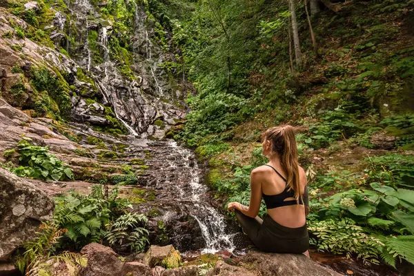 Ormandaki Pilj Dağı 'ndaki şelalenin yanındaki kayada oturan kadın turistin dikiz görüntüsü. Vahşi ormanda yalnız seyahat eden aktif bir dişi. Macera tatilleri, yaz yürüyüşleri doğa konsepti..