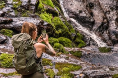 Pilj Şelalesi 'nin önündeki kayanın üzerinde duran sırt çantalı kadın turistin telefonuyla fotoğraf çekişini. Dağ ormanlarındaki kadın. Tek başına seyahat ve doğa konseptinde yürüyüş.