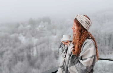 Kareli battaniyeye sarılı genç bir kadın balkon terasında duruyor, sisli kış gününde kahve içiyor. Örülmüş saç bandı takan kadın, bir fincan sıcak içecekle dondurucu dağ sabahının tadını çıkarıyor..